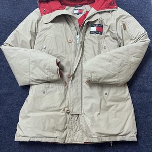 May include: A beige Tommy Hilfiger hooded jacket featuring a red interior lining, a front zipper closure with button toggles, and the Tommy Hilfiger logo patch on the left chest. The jacket includes long sleeves, a hood, and multiple pockets.