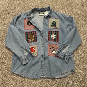 May include: A light blue denim long-sleeve button-down shirt featuring six embroidered holiday-themed patches on the front. The patches depict a wreath, a Christmas tree, a gingerbread man, a snowflake, a red mitten, and a snowman.