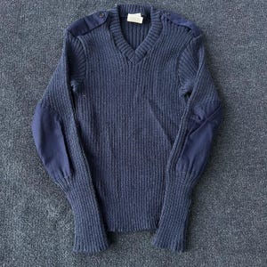 May include: A navy blue V-neck sweater featuring a ribbed knit texture, long sleeves, and solid fabric patches on the shoulders and elbows. The garment includes a small white label at the inner neckline and matching buttons on the shoulder epaulets.