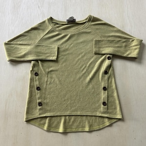 May include: Olive green long-sleeved shirt with a crew neck and side button details. The shirt has a rounded hemline and raglan sleeves. The buttons are dark brown. The fabric has a textured appearance.