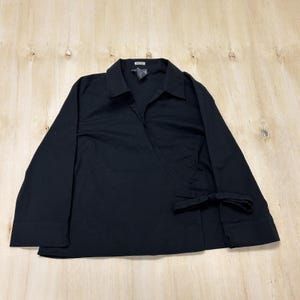 May include: A solid black long-sleeve wrap shirt featuring a pointed collar, a V-neckline, and a side tie closure. The garment has straight sleeves with cuffs and a hem that falls at the hip, constructed from a smooth, opaque fabric.