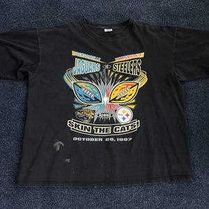 May include: A black short-sleeve t-shirt featuring a graphic for a 1997 Jacksonville Jaguars versus Pittsburgh Steelers football game. The design includes team names, logos, two footballs, and the text "SKIN THE CATS!" and "OCTOBER 26, 1997".