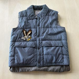 Könnte beinhalten: Eine schieferblaue, gesteppte Weste mit Umlegekragen, Druckknopfleiste auf der Vorderseite und zwei großen, rechteckigen aufgesetzten Taschen. Auf der Brust befindet sich ein gestickter Aufnäher eines Weißkopfseeadlers.