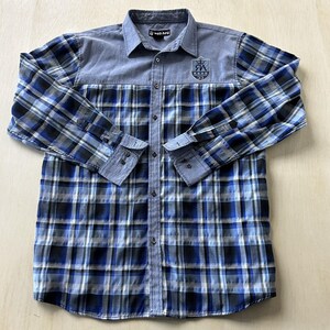 May include: A long-sleeved shirt with a blue and black check pattern on the lower half and sleeves. The upper section is a solid light blue. The shirt features a collar and a small emblem with text on the chest.