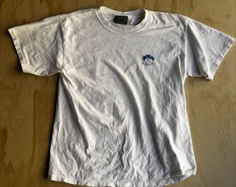 May include: Cream-coloured short-sleeved t-shirt featuring a small blue emblem on the chest. The emblem is a stylized palm tree design. The shirt has a crew neck and a tag at the back of the neck.