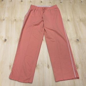May include: Coral-colored athletic pants with a drawstring waist and white side stripes. The pants have a relaxed fit and vertical seams down the front and back of the legs. The fabric appears to be a soft knit material.