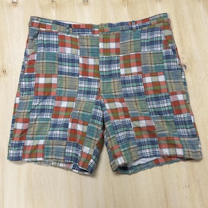 May include: A pair of shorts featuring a patchwork design made from various plaid fabric squares in shades of red, blue, green, and white. The shorts include a waistband with belt loops, a front button closure, and a zippered fly construction.