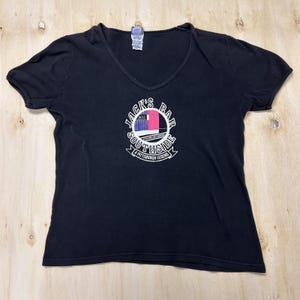 以下が含まれることがあります： 胸元に円形のグラフィックがプリントされた、黒の半袖VネックTシャツ。グラフィックには「JACK'S BAR SOUTHSIDE A PITTSBURGH LEGEND」の文字と、建物の外観を描いた小さな長方形の画像、および「12TH AND EAST CARSON」という文字が記されています。