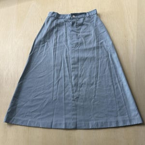 May include: A light blue A-line skirt featuring a structured waistband with belt loops, a single central button fastening, and a vertical front seam. The garment has a flared silhouette that extends to a straight hemline with visible fabric folds.