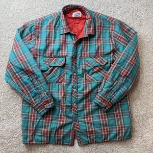 Puede incluir: Una camisa de manga larga con botones y un patrón de cuadros en tonos verde azulado, rojo y amarillo. La prenda incluye dos bolsillos en el pecho con cierre de botones, un cuello clásico y un forro interior rojo acolchado en el cuello.