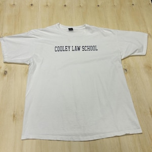 May include: A white t-shirt with the text "COOLEY LAW SCHOOL" printed in navy blue across the chest. The shirt is laid flat on a light-coloured wooden surface.