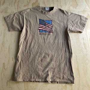 May include: A tan short-sleeve t-shirt featuring a graphic of a waving American flag inside a blue square. Below the flag, the text reads OKLAHOMA - U.S.A. The shirt has a crew neckline and a standard fit, with a visible interior neck label.