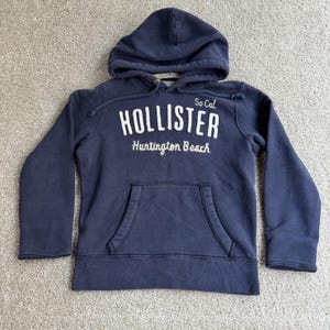 May include: A navy blue long-sleeve hooded sweatshirt featuring the white embroidered text "Hollister" and "Huntington Beach" with "So Cal" on the front. The garment includes a front kangaroo pocket, a drawstring hood, and ribbed cuffs.
