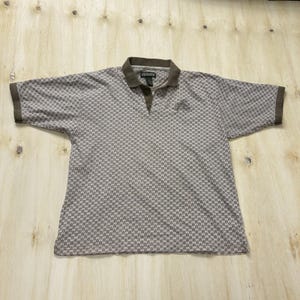 May include: A short-sleeve polo shirt in a light gray and brown color palette featuring a repeating horseshoe pattern. The garment has a solid brown collar, brown sleeve cuffs, and a three-button placket. An embroidered logo is on the chest.