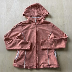 May include: A dusty rose zip-up hoodie featuring a full-length front zipper, a hood, and long sleeves. The garment includes white piping accents along the sleeves and torso, with ribbed white fabric panels visible at the lower side sections.