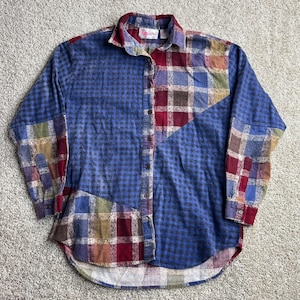 Könnte beinhalten: Ein langärmeliges Hemd mit Knopfleiste und einem Patchwork-Design aus blauem Vichy-Karo sowie mehrfarbigen Schottenmustern in Rot, Gelb und Grün. Das Hemd verfügt über einen klassischen Kragen, eine durchgehende Knopfleiste und einen abgerundeten Saum.