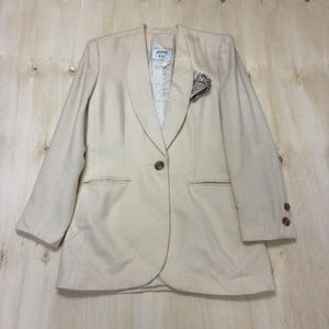 May include: A cream-colored blazer with a notched lapel, a single button closure, and two front pockets. The blazer has a pocket square and two buttons on each sleeve. The tag reads "Gasper A.S.L."