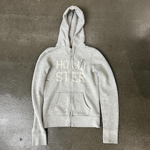 May include: A light gray zip-up hoodie featuring a hood with drawstrings, long sleeves, and two front pockets. The word HOLLISTER is embroidered in white across the chest. The garment is constructed from a soft, heathered fabric material.