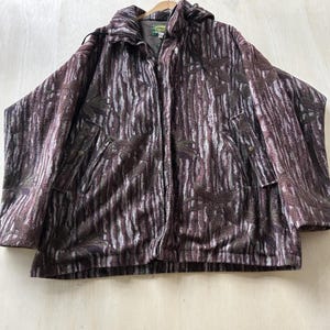 Puede incluir: Una chaqueta de forro polar de manga larga con un patrón de camuflaje de corteza de árbol en tonos marrón y gris con siluetas de hojas oscuras. La chaqueta incluye cierre de cremallera frontal, capucha con cordones y dos bolsillos laterales con botones a presión.
