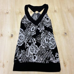 May include: Black and white patterned tank top with a V-neck and halter straps. The top features a floral and paisley design. The bottom hem is solid black, and the straps connect at the back with a ring detail.