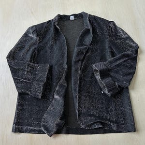 May include: A black open-front blazer featuring a subtle metallic silver shimmer throughout the fabric. The garment has three-quarter length sleeves with folded cuffs, a simple collarless neckline, and a straight hemline with a textured finish.