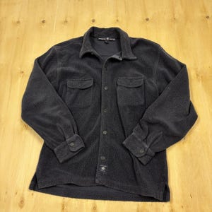 Könnte beinhalten: Ein dunkelgraues Fleece-Hemd mit Knopfleiste, zwei Brusttaschen und geknöpften Manschetten. Das Hemd hat einen Kragen und ein kleines Logo unten. Der Stoff wirkt weich und strukturiert.