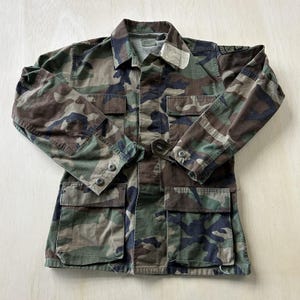 May include: A long-sleeve camouflage jacket featuring a woodland pattern in shades of green, brown, and black. The jacket includes a pointed collar, a button-front closure, two chest pockets with flaps, two lower pockets with flaps, and buttoned cuffs.