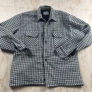 May include: A long-sleeve button-up shirt featuring a black and white checkered pattern. The shirt includes a collar, a front button closure, and two chest pockets with buttoned flaps. An Ozark Trail brand label is visible on the inner neck.