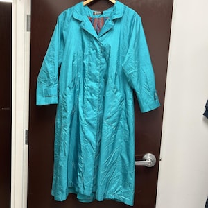 Può includere: Un lungo cappotto turchese brillante con colletto ampio, maniche lunghe con polsini abbottonati e due grandi tasche frontali. La fodera interna presenta un vivace motivo a righe verticali multicolori nei toni del rosso, blu e giallo.