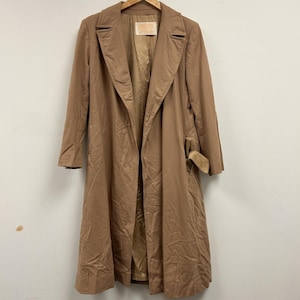 May include: A long, tan-colored trench coat featuring wide lapels, long sleeves, and a matching fabric belt tied at the side. The coat has a visible interior lining and a brand label sewn into the inner neckline area, presenting a classic silhouette.