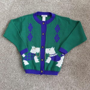 May include: A green knit cardigan featuring a purple collar, cuffs, and button placket. The sweater displays two white terrier dogs, purple argyle patterns, and gold-toned chain embellishments. It has five buttons and a white interior neck label.