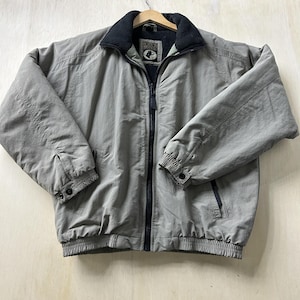 May include: A light gray jacket with a black collar and a full-length zipper. The jacket has two front pockets and elastic cuffs. The brand logo is visible on the inside of the jacket. The jacket is hanging on a wooden hanger.