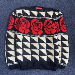 May include: A long-sleeve knit sweater featuring a black and white geometric triangle pattern on the lower section and sleeves. The upper chest area displays a horizontal band of red rose motifs against a black background with a red neck label.