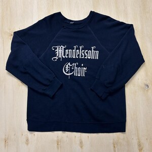 Könnte beinhalten: Ein dunkelblaues Langarm-Sweatshirt mit dem in weißer, gotischer Schrift aufgedruckten Text Mendelssohn Choir. Das Kleidungsstück verfügt über einen Rundhalsausschnitt, Raglanärmel sowie gerippte Bündchen an den Ärmeln und am Saum.