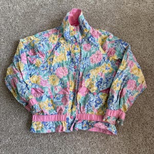 May include: A long-sleeve jacket featuring a floral pattern in shades of pink, yellow, and blue. The jacket has a high collar, a front button closure, pink elasticized cuffs, and a pink elasticized waistband with matching pink fabric accents.