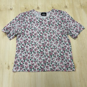 May include: White short-sleeved t-shirt with a pattern of small pink roses and green leaves. The shirt has a crew neck and the brand name "FRITZ" is printed on a small black tag at the neckline.
