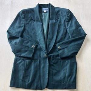 Puede incluir: Un blazer verde azulado oscuro con cierre de un solo botón, dos bolsillos delanteros y puños abotonados. El blazer tiene un diseño de solapa clásico y un corte entallado. El forro interior es de un tono verde azulado más claro.