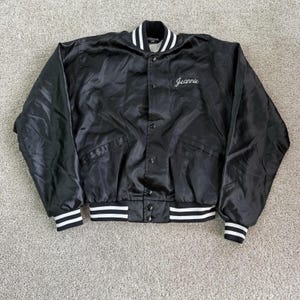 May include: Black satin bomber jacket with white and black striped cuffs and collar. The jacket has a button-up front and two front pockets. The name "Jeannie" is embroidered in white script on the chest.