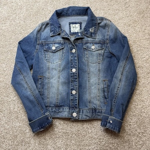 May include: A blue denim jacket with a button-up front, two chest pockets, and two side pockets. The jacket has a classic collar and buttoned cuffs. The brand tag reads "Mudd".
