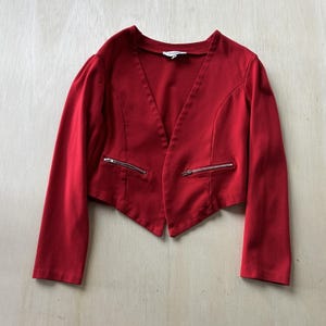 以下が含まれることがあります： Vネックのデザインと長袖の赤いブレザー。シルバーのジッパーが付いた2つのフロントポケットがあります。この衣服は無地の赤い生地で作られています。