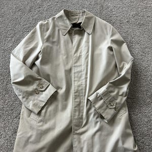 May include: A beige trench coat featuring a classic collar, a concealed button placket, and long sleeves with adjustable buttoned cuffs. The coat is constructed from a smooth, solid-colored fabric and includes a structured collar design.