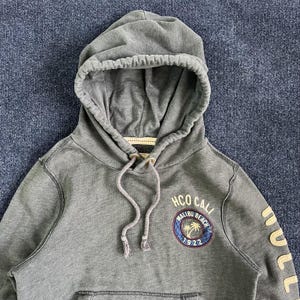 May include: A charcoal gray hooded sweatshirt featuring a circular patch on the chest with the text HCO CALI MALIBU BEACH 1922 and a palm tree graphic. The hoodie includes a drawstring with frayed ends and light-colored lettering on the sleeve.