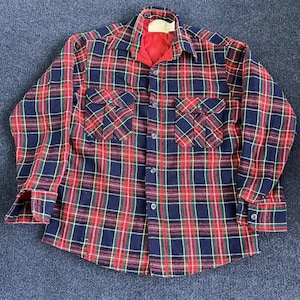 May include: A long-sleeve button-up shirt featuring a plaid pattern in navy blue, red, and yellow. The shirt has a pointed collar, two chest pockets with buttoned flaps, and a red quilted interior lining visible at the neckline and front opening.