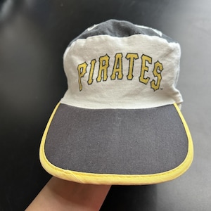 Puede incluir: Una gorra de béisbol con un panel frontal blanco que presenta la palabra PIRATES impresa en amarillo con bordes negros. La gorra tiene una corona gris oscuro y una visera a juego con un borde amarillo en el contorno.