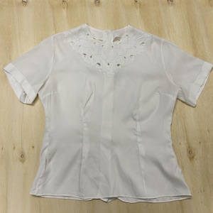 May include: A short-sleeved white blouse featuring a round neckline with intricate white cutwork embroidery. The garment has a button-down back closure, cuffed sleeves, and vertical seam detailing down the front, providing a structured fit.
