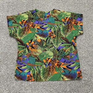 May include: A short-sleeved blouse featuring a vibrant botanical print with large green leaves, bright orange flowers, and purple accents. The garment has a round neckline and a relaxed fit, showcasing a dense, colorful pattern of tropical foliage.