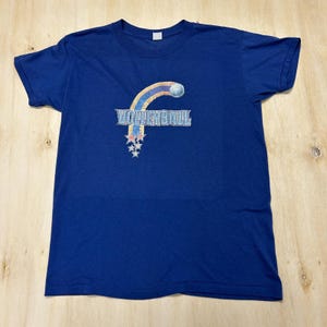 May include: A royal blue short-sleeve t-shirt featuring a graphic on the chest. The design includes the word VOLLEYBALL in a light blue textured font, arched by a curved rainbow-colored stripe with a volleyball and several small stars.