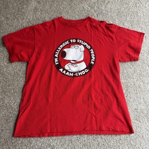 May include: A bright red short-sleeved t-shirt featuring a circular graphic in the centre. The graphic depicts a white cartoon dog with a red collar and crossed arms, surrounded by the text I'M ALLERGIC TO STUPID PEOPLE AAAH-CHOO.