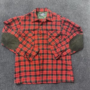 May include: A long-sleeve button-down shirt featuring a red and black plaid pattern, a pointed collar, two chest pockets with flaps, and dark green suede elbow patches. The interior collar label reads Pendleton, and the inner lining is green.
