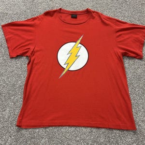 May include: A bright red short-sleeved t-shirt featuring a central graphic of a yellow lightning bolt inside a white circle with a thin black outline. The shirt has a crew neckline and a standard fit, displaying a classic superhero-inspired design.